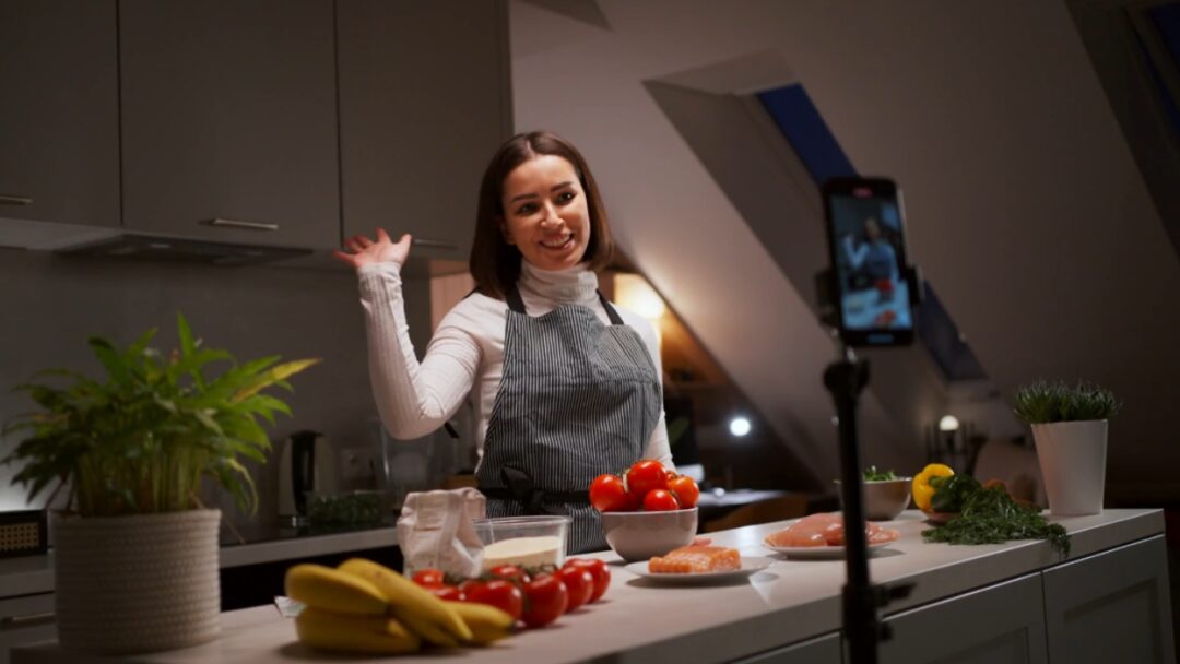 A woman records an online culinary lesson beside fresh food on a kitchen counter